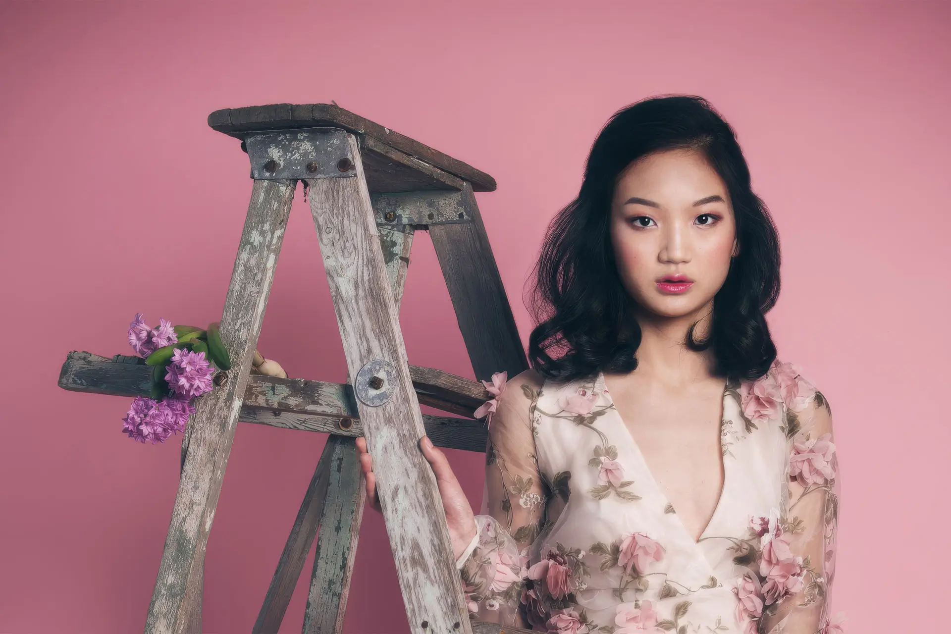 A woman in a floral dress stands beside a ladder, smiling and looking towards the camera in a bright indoor setting.