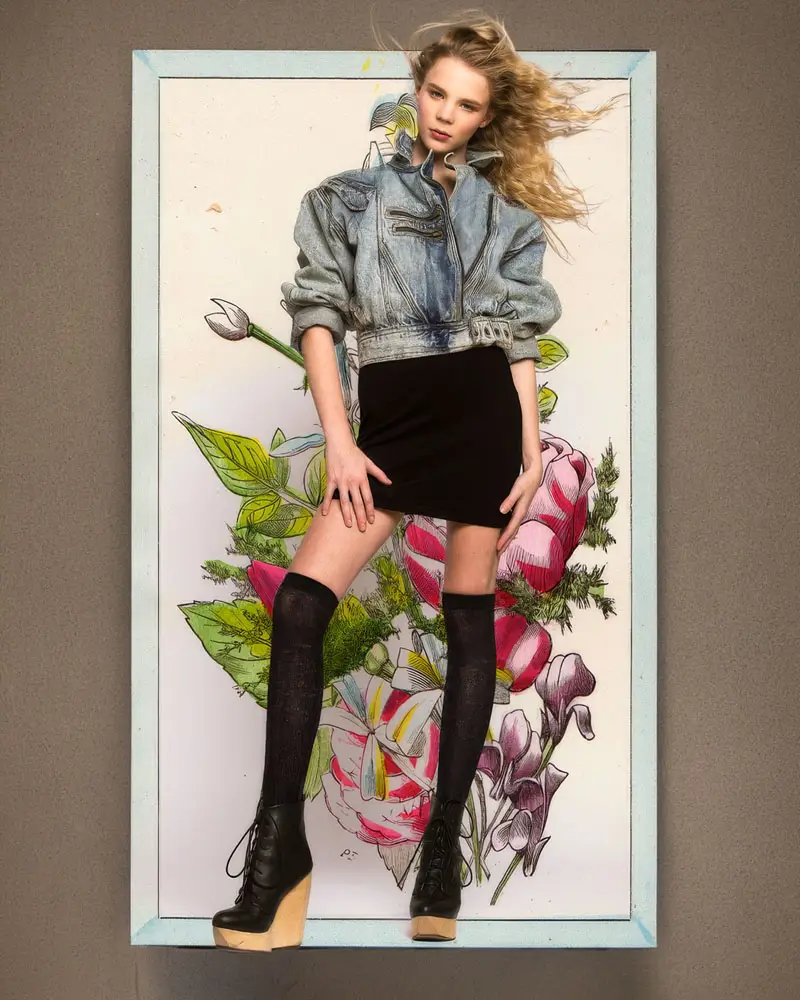 A woman wearing a denim jacket and a floral skirt stands in front of a painting depicting greenery.