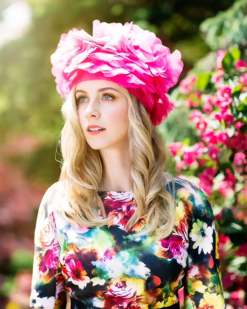 A beautiful blonde woman in a floral dress and a bright pink hat, smiling against a vibrant outdoor background.