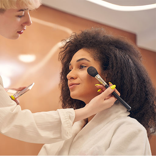 A woman applies makeup to another woman, who is seated and looking in a mirror, in a well-lit beauty studio.